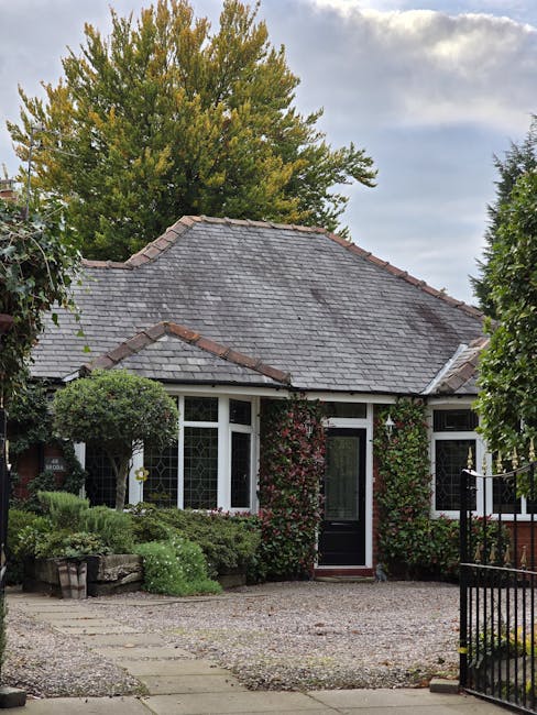 A small front garden in front of a detached house with a slate-tiled roof and white-framed windows. The garden features neatly trimmed low hedges and leafy green bushes along the front of the house, w