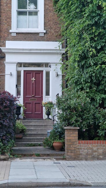 A small front garden in front of a detached house with a slate-tiled roof and white-framed windows. The garden features neatly trimmed low hedges and leafy green bushes along the front of the house, w